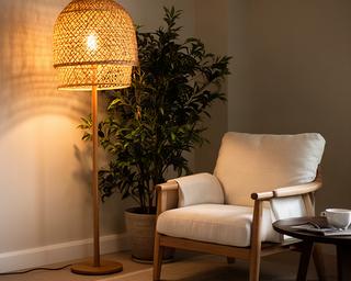 Lámpara de pie con pantalla de ratán y luz cálida junto a una planta de interior y un sillón de lectura